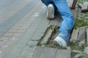 A person wearing jeans and white shoes is kneeling on a sidewalk with one leg in a hole beside a damaged curb.