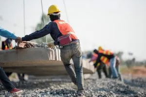 Construction workers in safety gear guide a large concrete slab being lifted by a crane at an outdoor worksite.