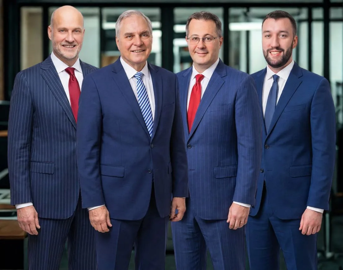 Four men in suits stand side by side in an office setting, looking at the camera and smiling.