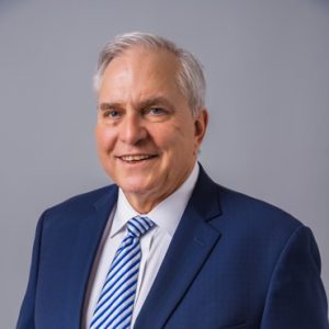 Smiling older man with gray hair wearing a blue suit, white shirt, and blue striped tie, posing against a plain gray background.