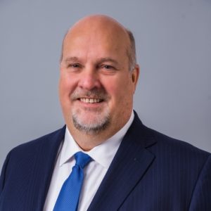 Bald man with a goatee wearing a dark blue suit, white shirt, and blue tie, smiling in front of a plain gray background.