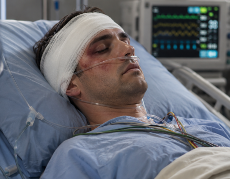 A man with a head injury and facial bruising lies unconscious in a hospital bed, connected to medical monitors and tubes.
