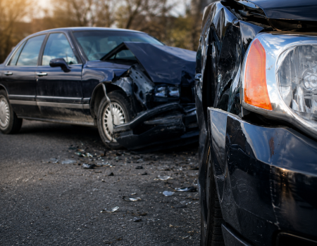 Two dark-colored cars with visible front-end damage are involved in a collision on a street, with debris scattered on the ground.