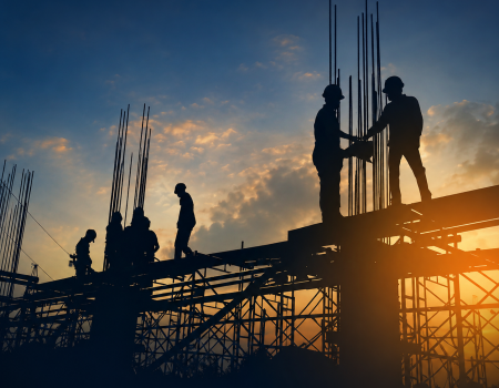 Silhouetted construction workers stand on scaffolding at a building site during sunset, with rods and beams visible against the evening sky.