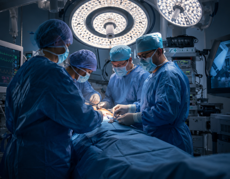 Four surgeons in blue scrubs and masks perform an operation under bright surgical lights in a modern operating room with medical equipment and monitors.