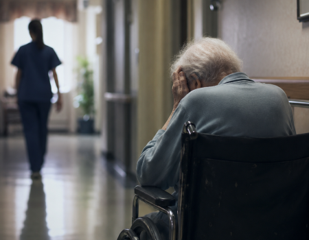 An elderly person in a wheelchair sits in a hallway with their head in their hands while a healthcare worker walks away in the background.