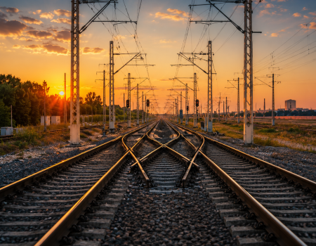 Railway tracks converging and diverging under overhead wires at sunset, with poles and signals visible, and an orange sky in the background.